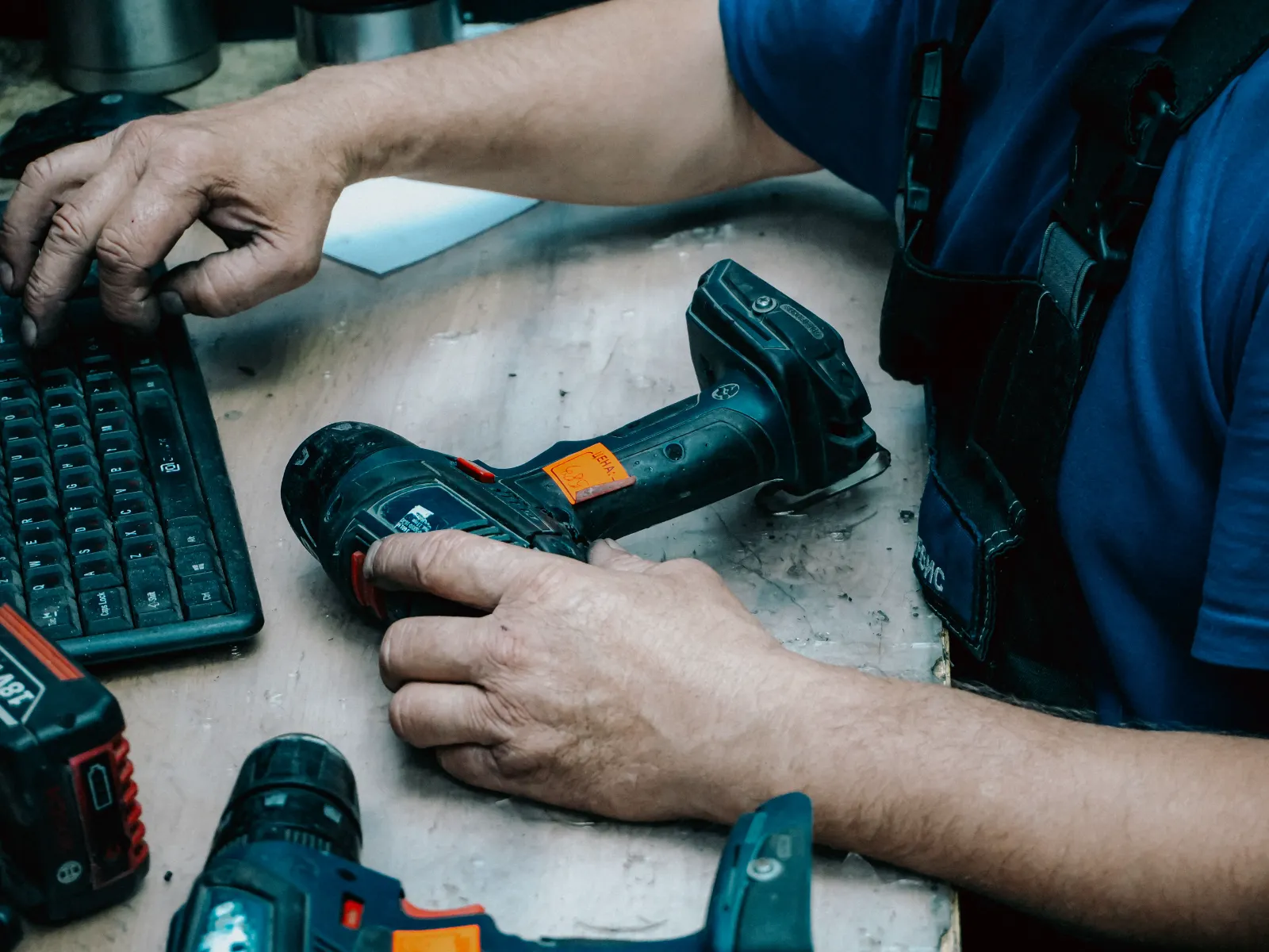 A worker's hands on a cordless drill beside a keyboard on a workbench